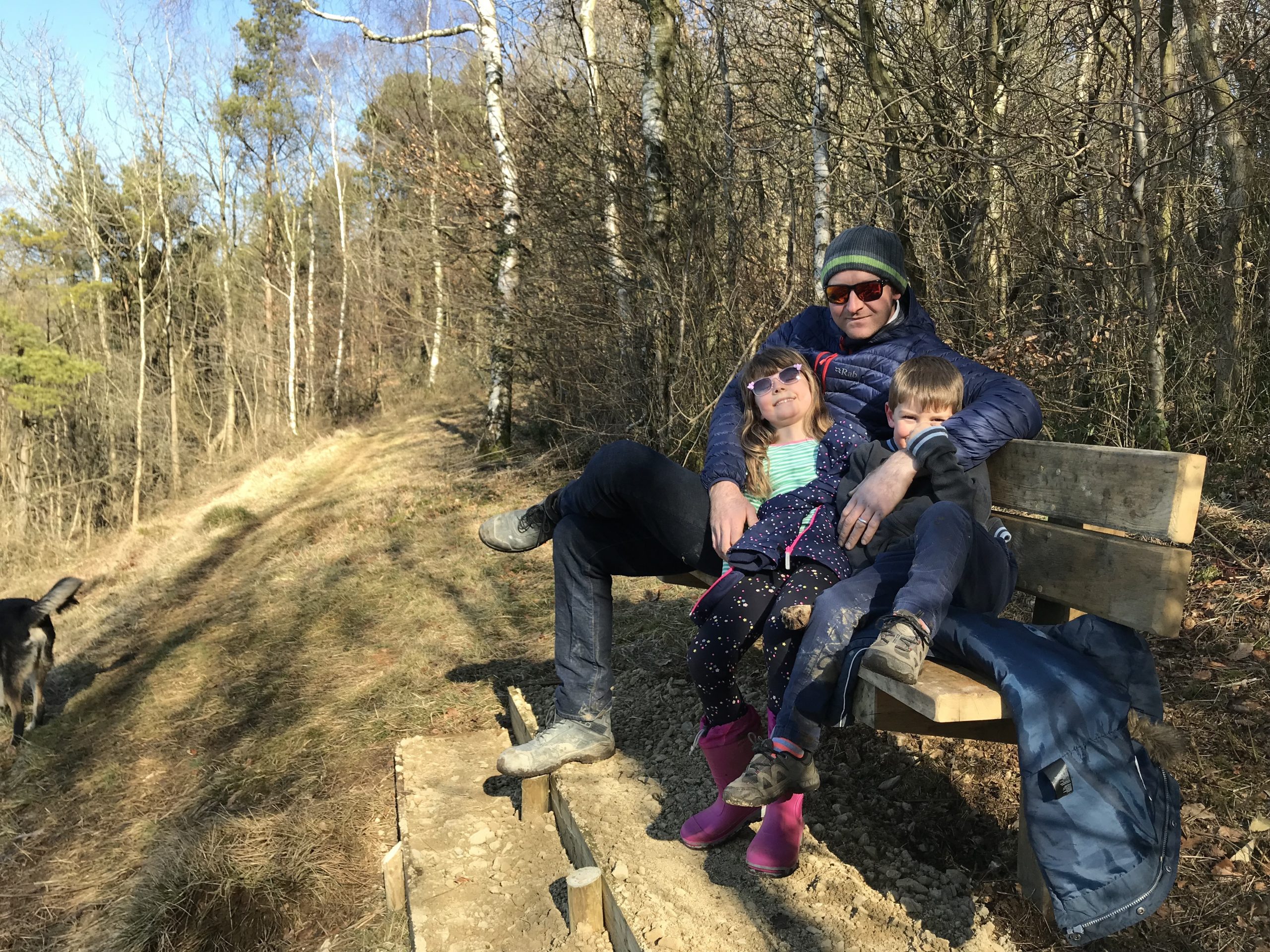 My husband and two children cuddled up on a bench at a viewpoint in a lovely English wood with a blue sky above them, wrapped up warm but wearing sunglasses.